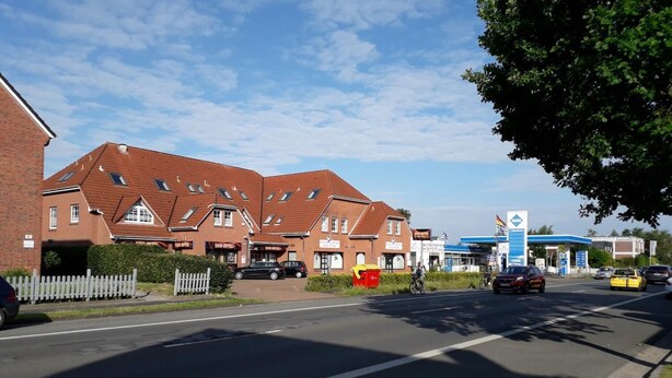 A row of red-brick houses with pitched roofs, a gas station, and a cyclist on the road. - Pension Norddeicher Straße (Norden)