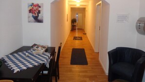 A hallway with a wooden floor, a dining area with a table and chairs, a black armchair, and a painting on the wall. - Pension Norddeicher Straße (Norden)