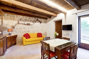 A cozy living room with a stone wall, wooden beams, a yellow sofa, a dining table, and a TV mounted on the wall. - Authentic Villa surrounded by Nature CIN IT055032C29Z019674 (Terni, Fraz. Torre Orsina)