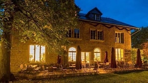 A stone building with a pitched roof, large windows, and a well-lit outdoor area with chairs and umbrellas. - Hotel Menschels Vitalresort (Meddersheim)