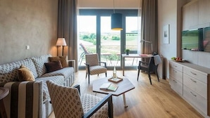 A modern living room with a sofa, armchair, coffee table, and a dining area with a table and chairs. There is a large window providing a view of the outdoors. - Hotel Menschels Vitalresort (Meddersheim)