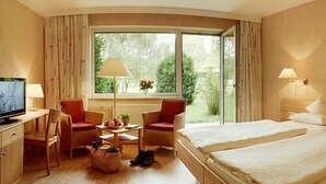 A hotel room with a bed, two chairs, a small table, a TV, and a window with curtains. - Hotel Menschels Vitalresort (Meddersheim)