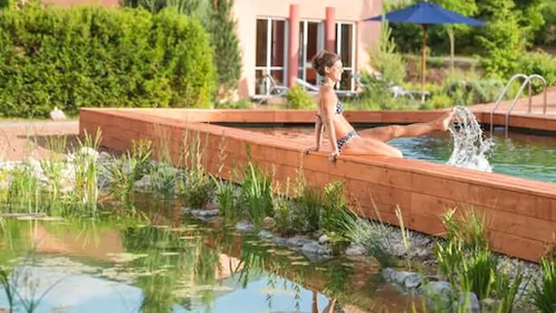 A swimming pool with a wooden deck, surrounded by greenery and a building in the background. - Hotel Menschels Vitalresort (Meddersheim)