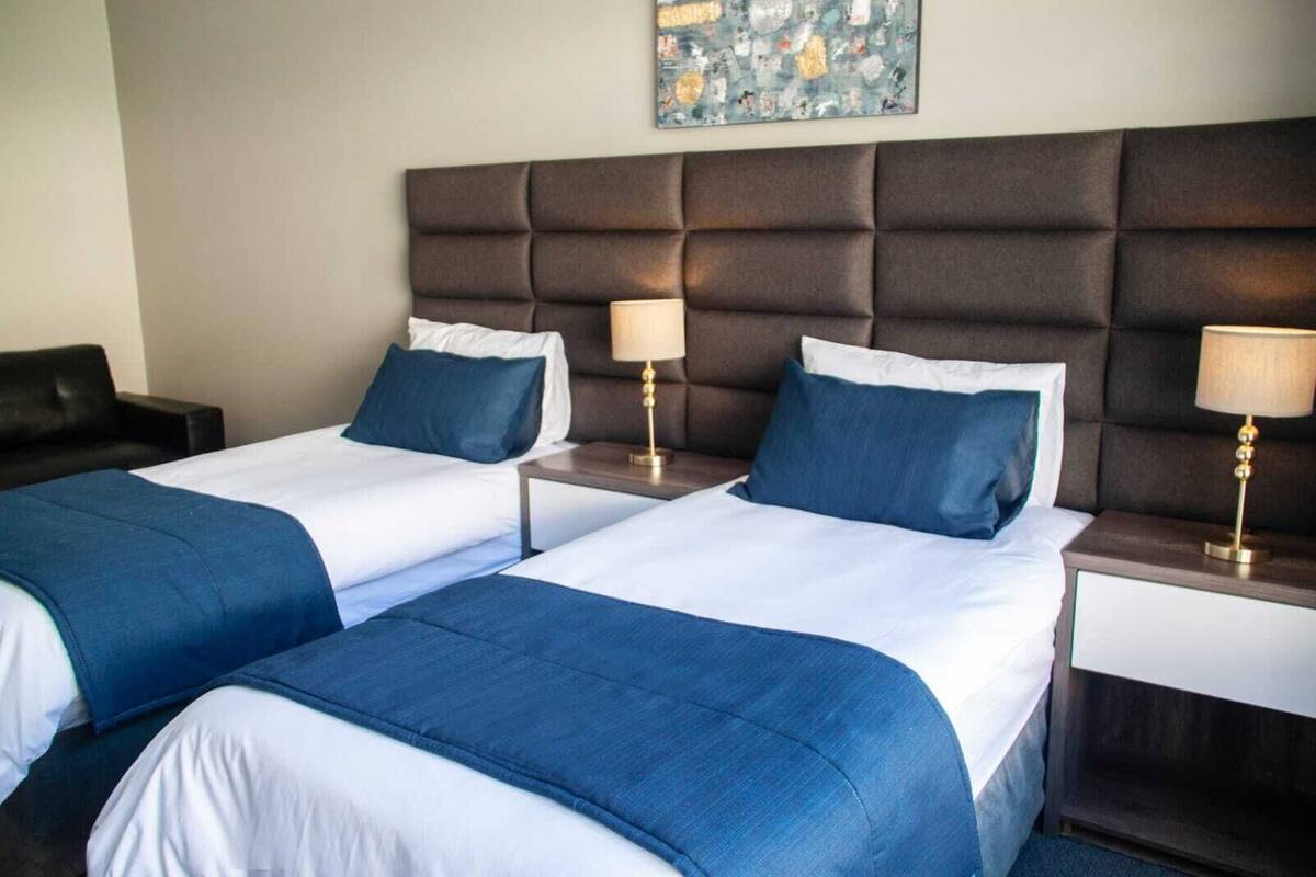 A hotel room with two beds, each with a blue bedspread and white linens. There are two bedside tables with lamps.