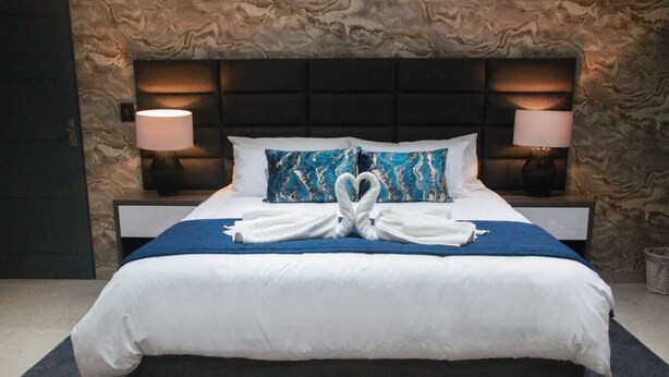 A neatly made bed with white linens, two pillows with a blue and white pattern, and two white towels folded into swans on a blue bedspread. - The Cato Suites Hotel (Durban)