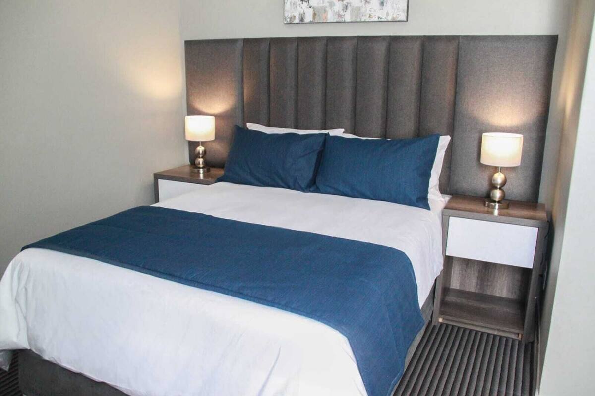 A neatly made bed with a white and blue bedspread, flanked by two bedside tables with lamps, in a room with a grey padded headboard and a framed picture above the bed.