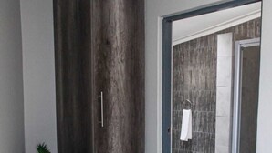 A hallway with a dark wood wardrobe, a small plant on the left, and a tiled shower area in the background. - The Cato Suites Hotel (Durban)