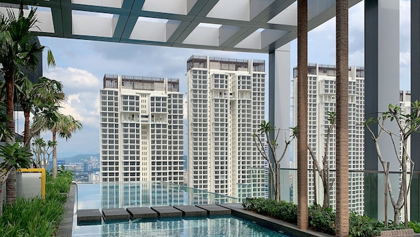A rooftop pool area with a glass table and wicker chairs, surrounded by tall buildings and greenery.