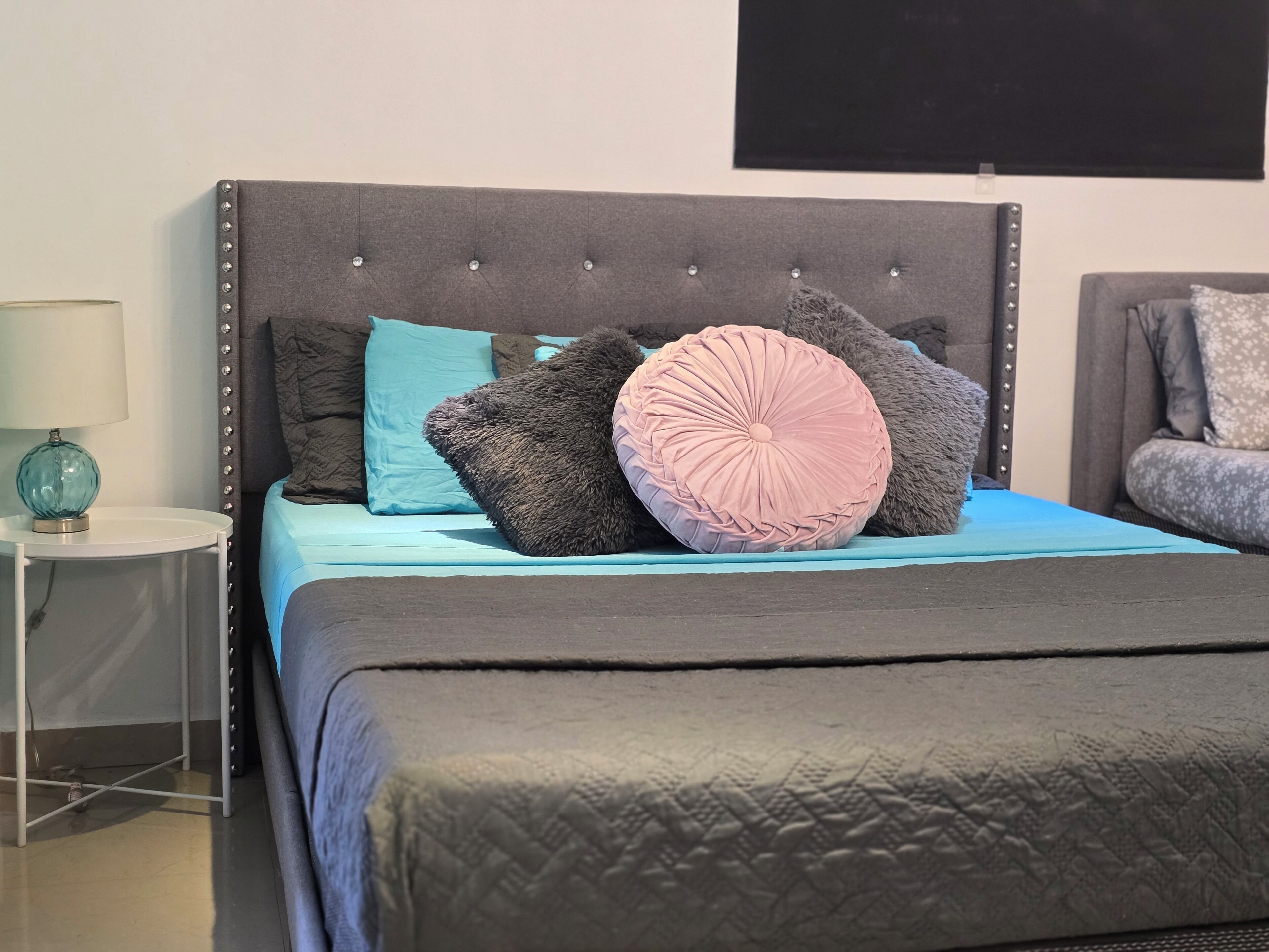 Een bed met een gestoffeerd hoofdbord, een blauwe spreideken en meerdere kussens.