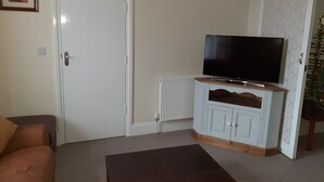 A living room with a white door, a television on a wooden cabinet, a brown sofa, and a radiator. - Family holiday home near both sea front and town (Yarmouth)