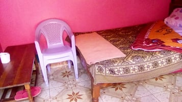 A room with a wooden table, a plastic chair, a bed with a patterned bedspread, and a tiled floor.