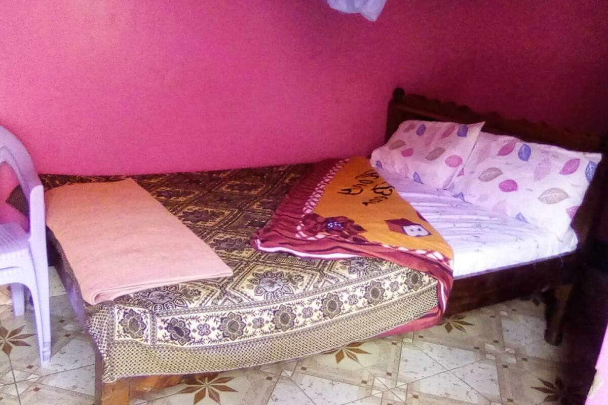 A small, single-bed room with a patterned bedspread, a pink chair, and a patterned rug on a tiled floor.