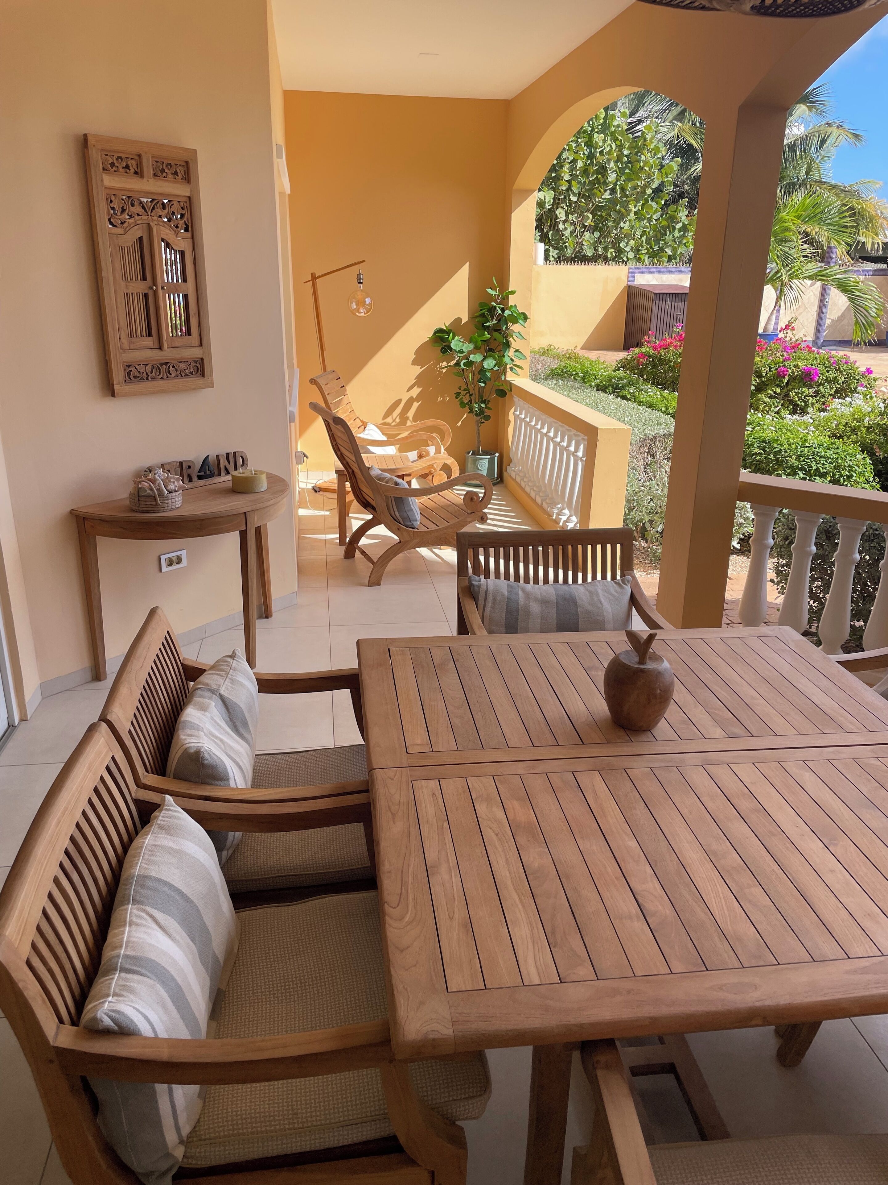 Een houten zitgedeelte buiten met een tafel en banken, een decoratief wandobject en uitzicht op een tuin met planten en bloemen.