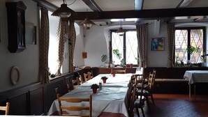 A dining area with long tables, wooden chairs, and hanging lamps. There are potted plants and a painting on the walls. - Haus Barnabas im Engel - Gasthaus Engel (Utzenfeld)