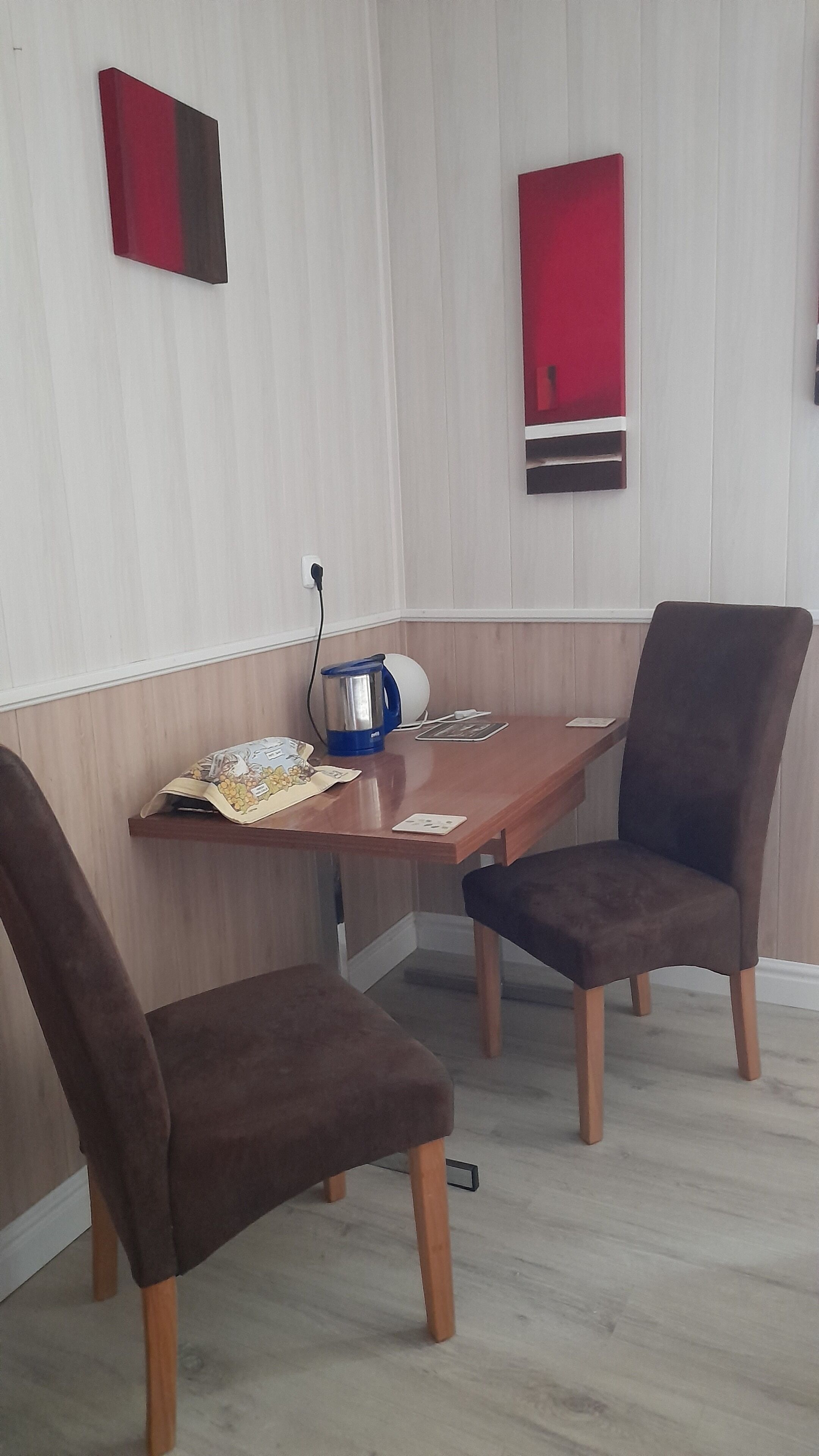 Un comedor con una mesa de madera, dos sillas marrones, un hervidor azul y un adorno rojo en la pared.