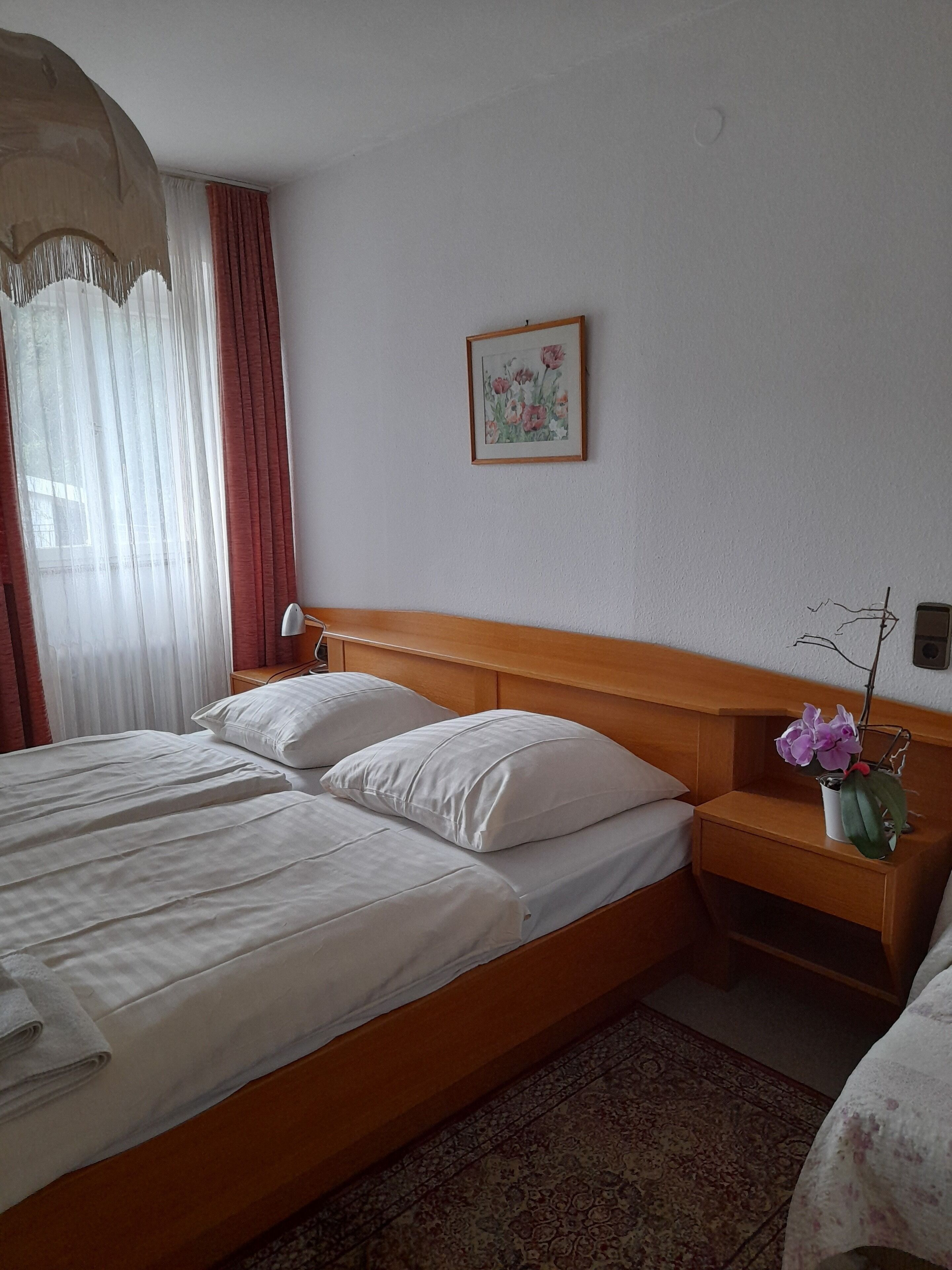 Un cuarto de hotel con una cama grande, una mesita de noche con lámpara, un jarrón pequeño con flores y un cuadro en la pared.