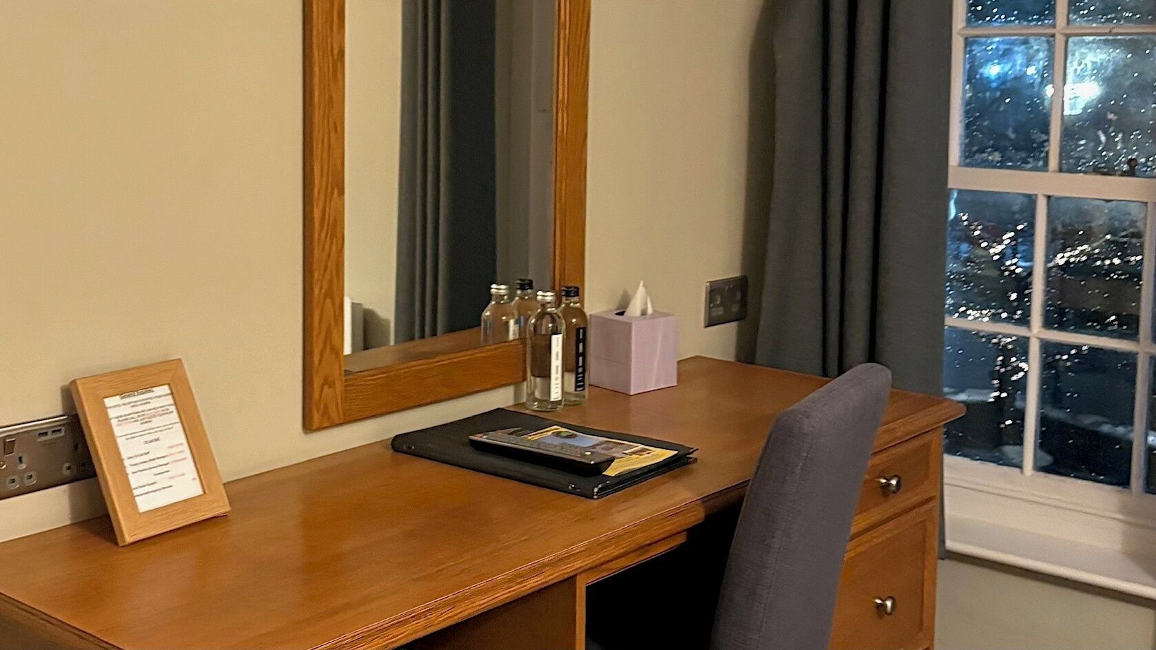 A wooden desk with a mirror, a framed menu, a tissue box, and a bottle of hand sanitizer.