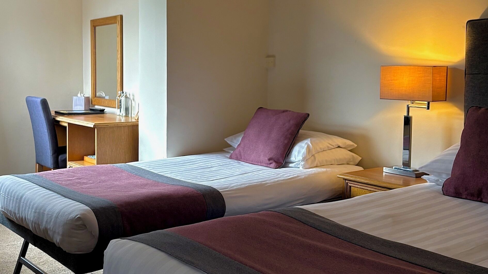 A hotel room with two beds, a desk, a chair, and a lamp.