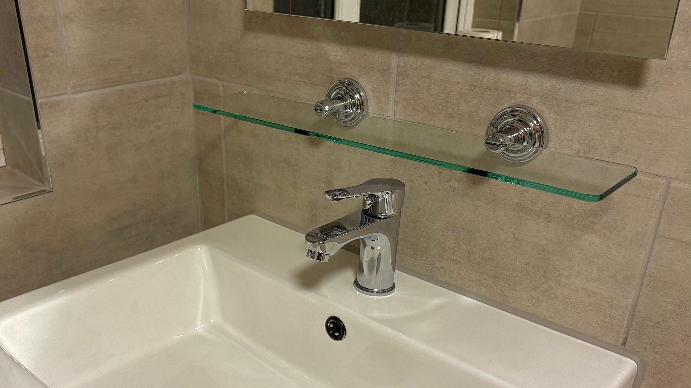 Un baño con un lavamanos blanco, grifo cromado y un estante de vidrio arriba.