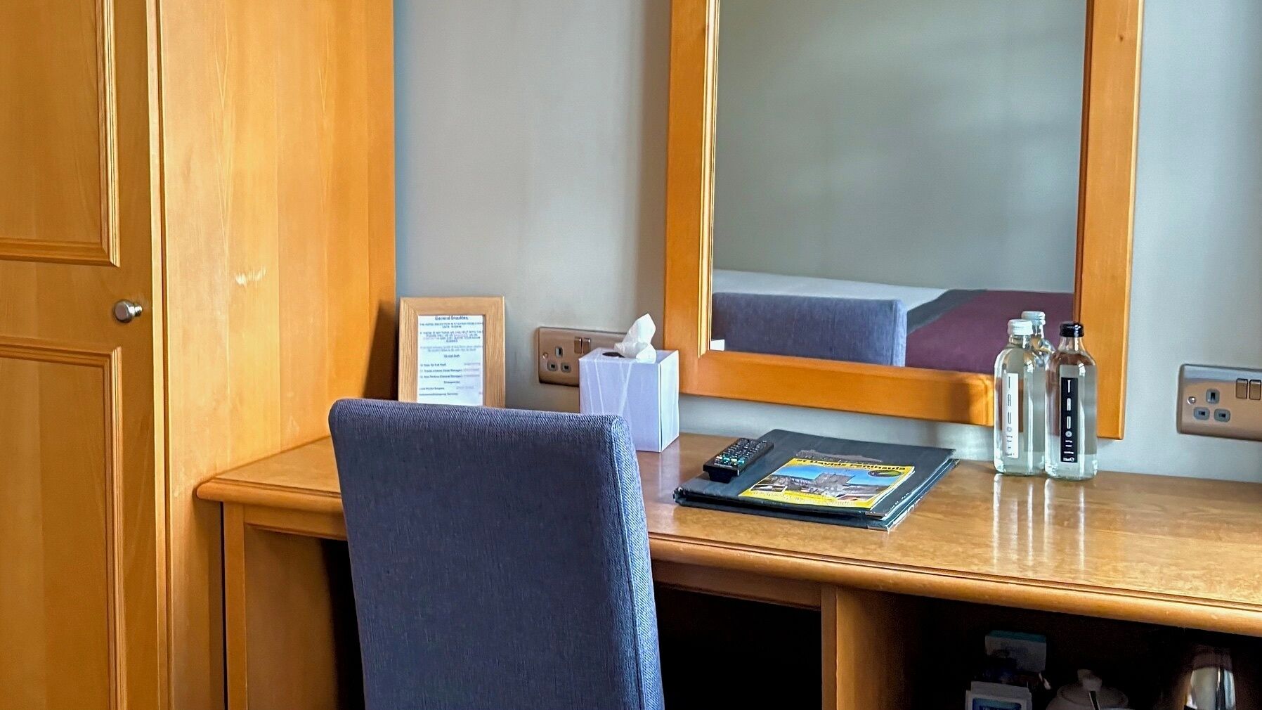 Camera d'albergo con una scrivania in legno, una sedia, uno specchio, un telecomando, una rivista, un dispenser di fazzoletti e alcune bottiglie.