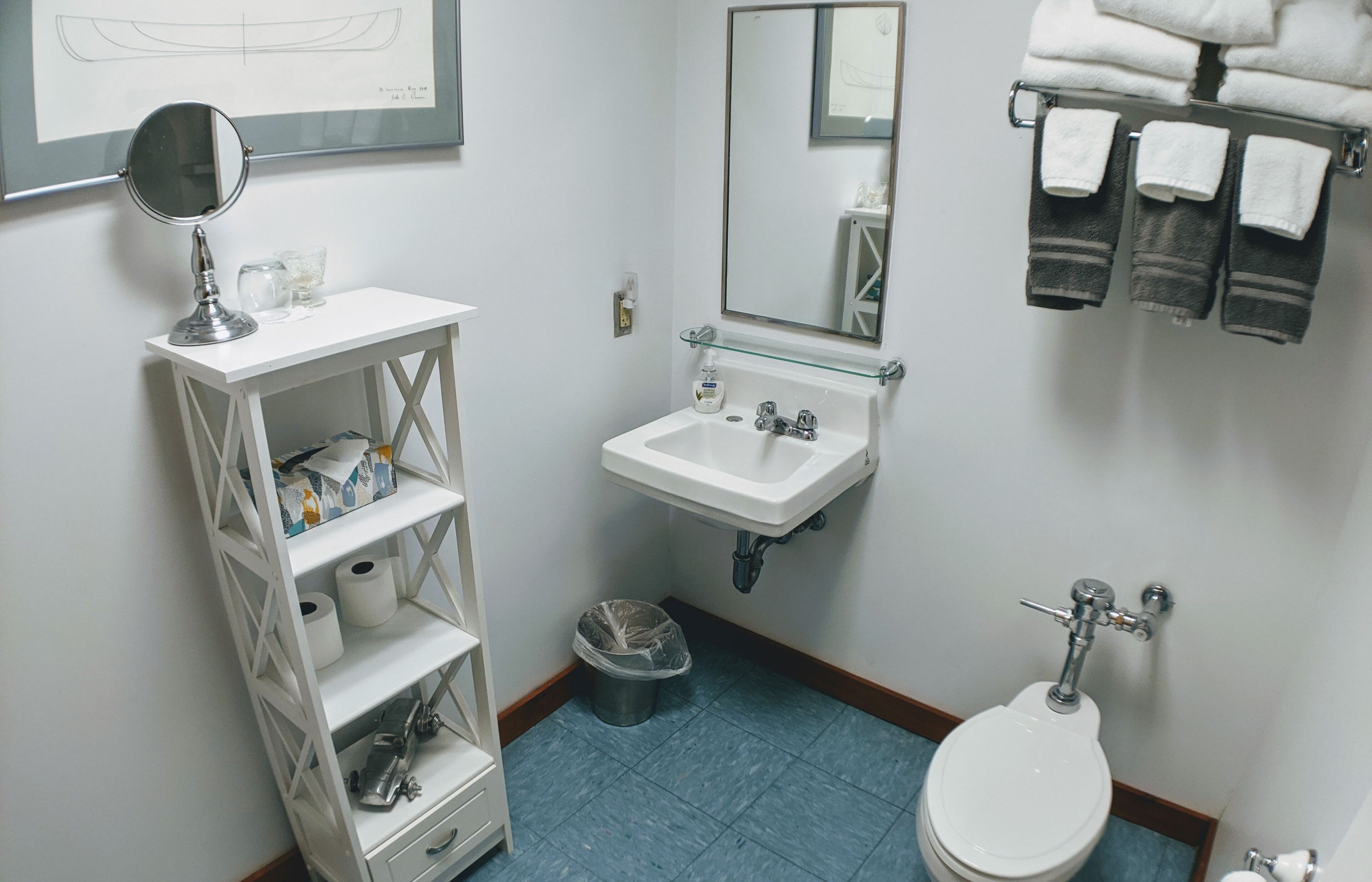 Et badeværelse med hvid vask, spejl, toilet og et hyldesæt med toiletartikler.