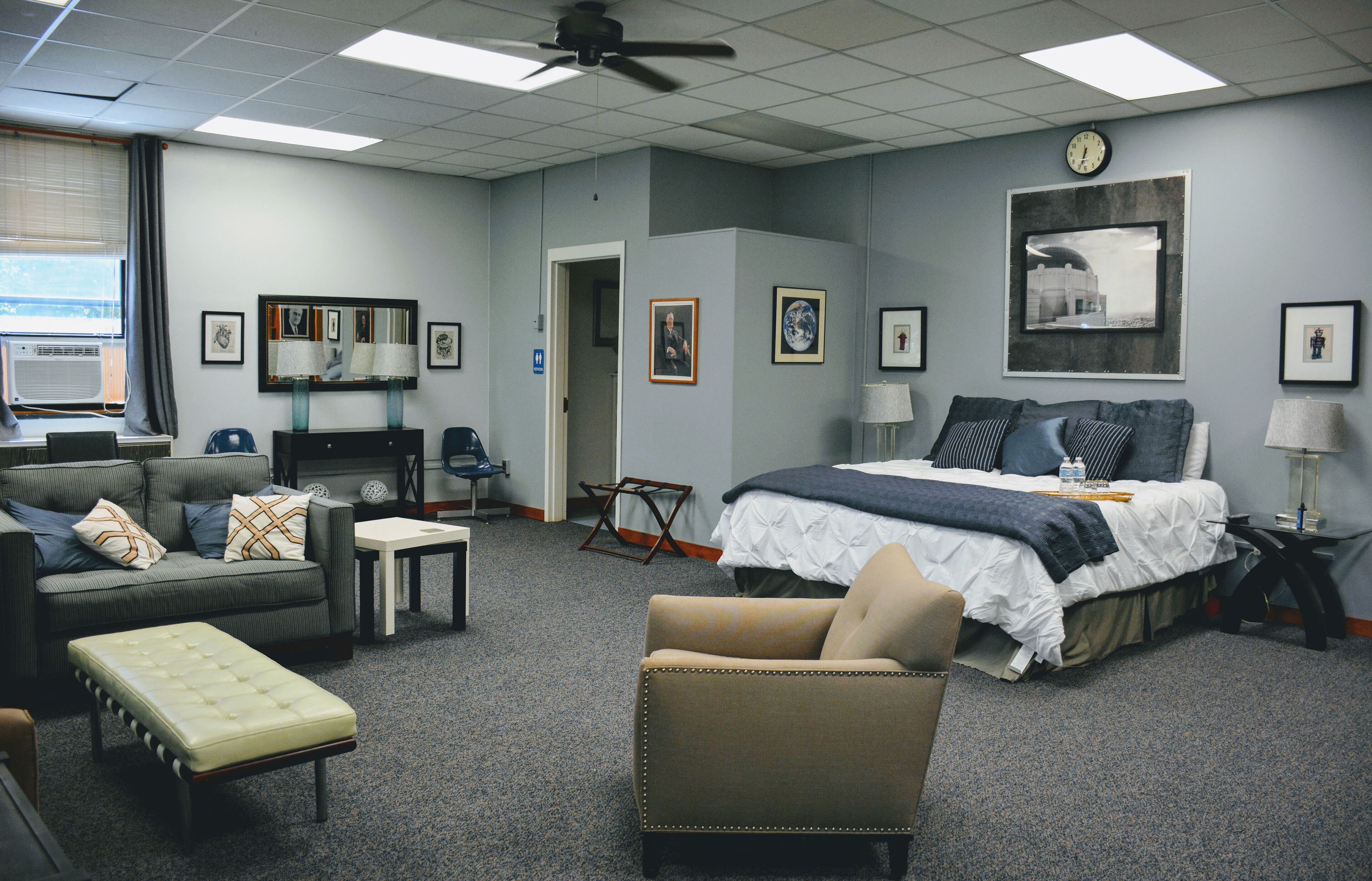 A hotel room with a bed, a sofa, a chair, a small table, and a desk. There is a window with blinds, a ceiling fan, and a clock on the wall.