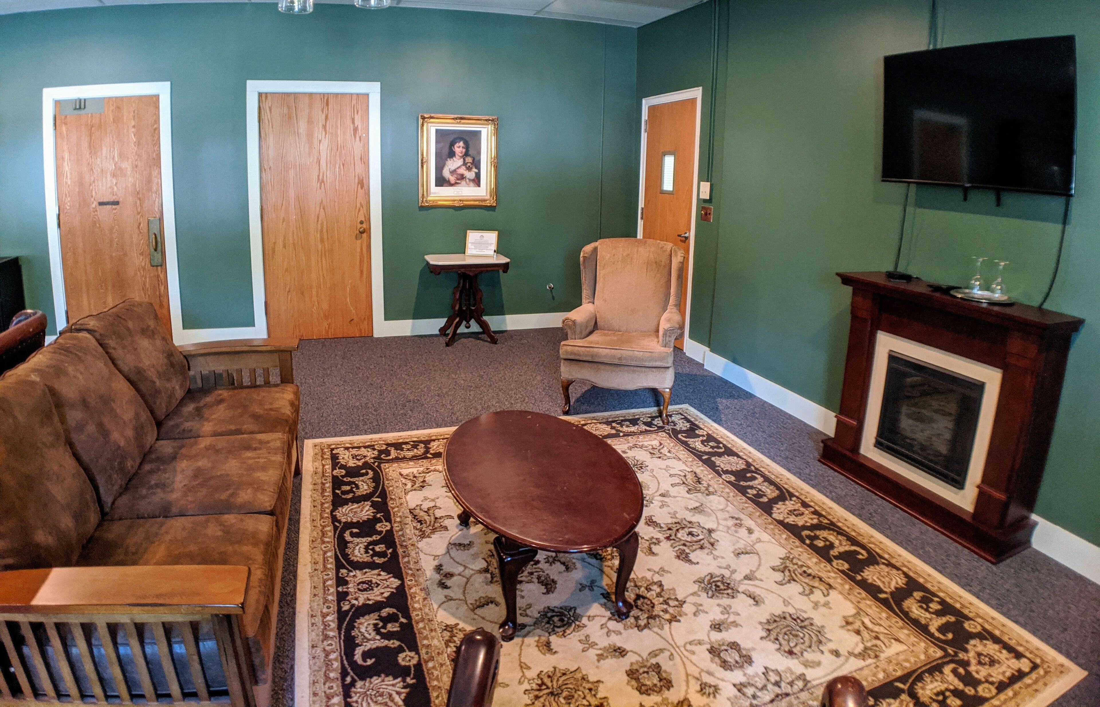A living room with a brown sofa, a round table, a patterned rug, a wooden chair, a small table with a framed picture, and a fireplace.