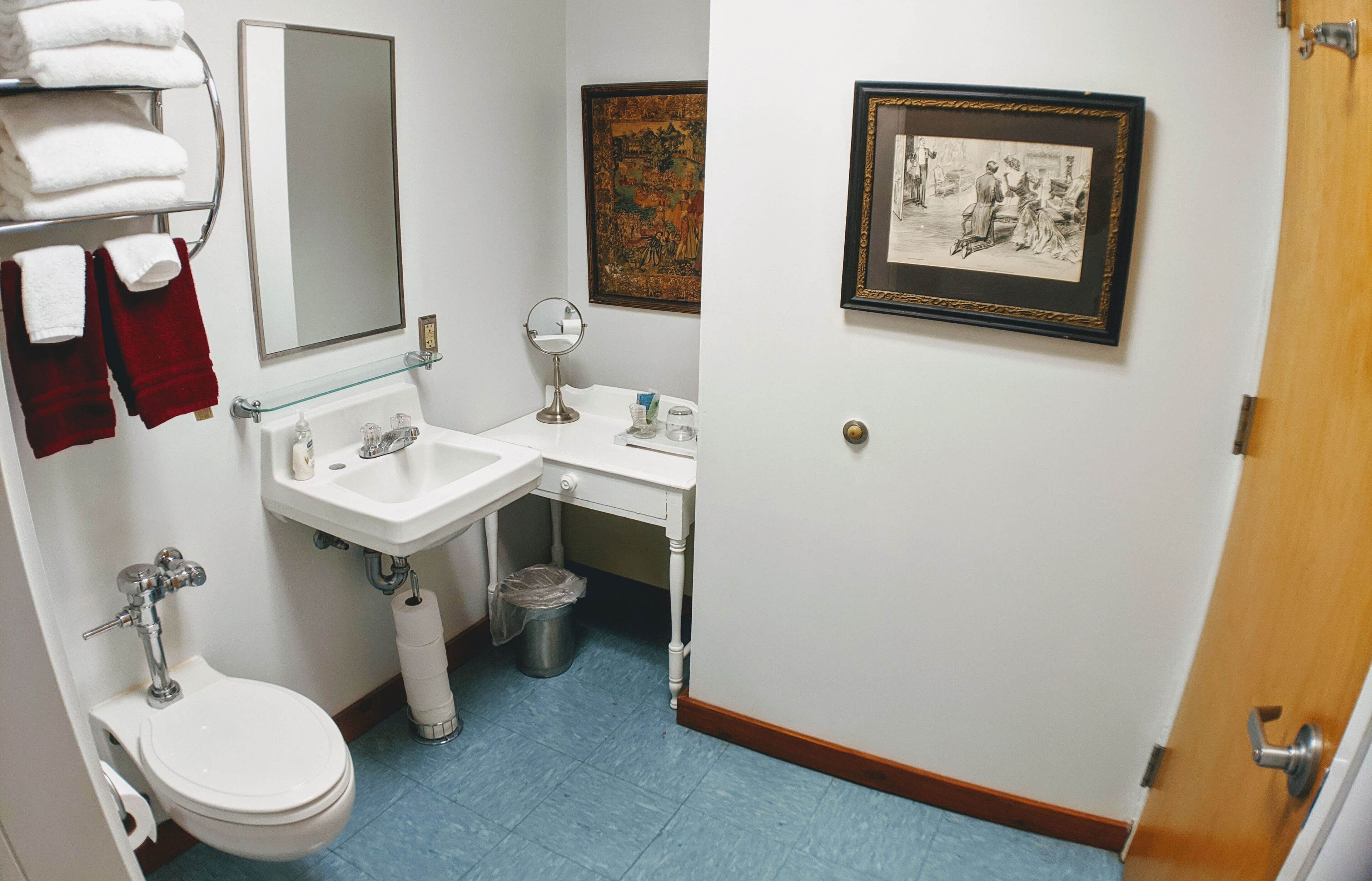 Une salle de bain avec des toilettes, un lavabo et un miroir. On y trouve des serviettes sur un porte-serviettes, une poubelle et deux tableaux encadrés accrochés au mur.