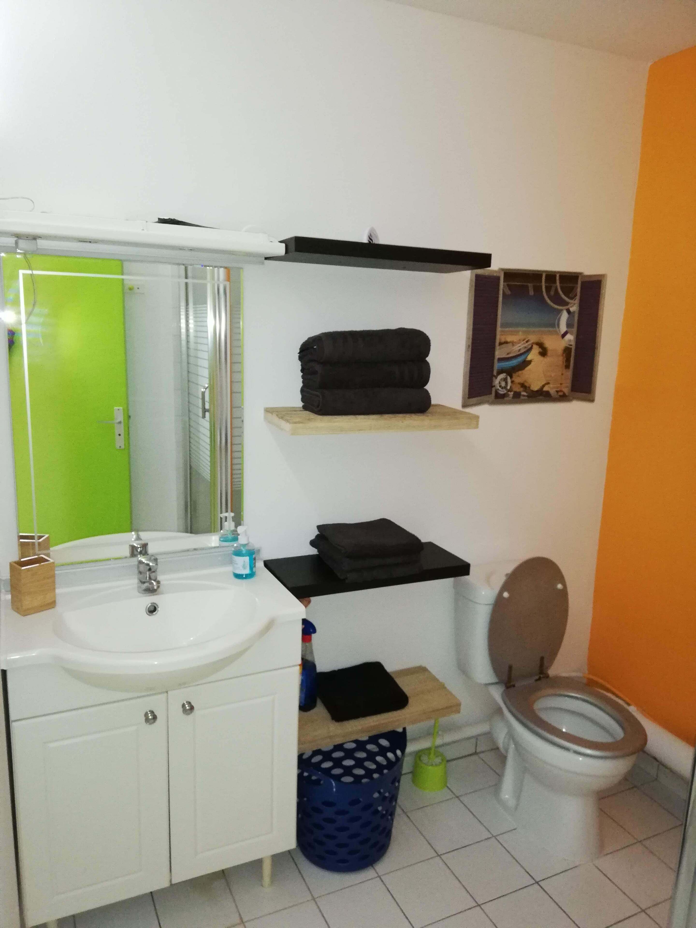 A bathroom with a white sink, mirror, and toilet. There are shelves with towels and a blue trash bin.