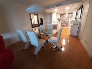 A dining area with a glass table and white chairs, a kitchen with a stone backsplash, and a red chair in the corner.