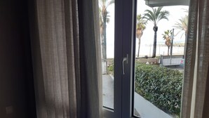 A view through a window with a curtain partially drawn, revealing a balcony with a hedge and palm trees. - Sunset on the beach, Salou (Tarragona)