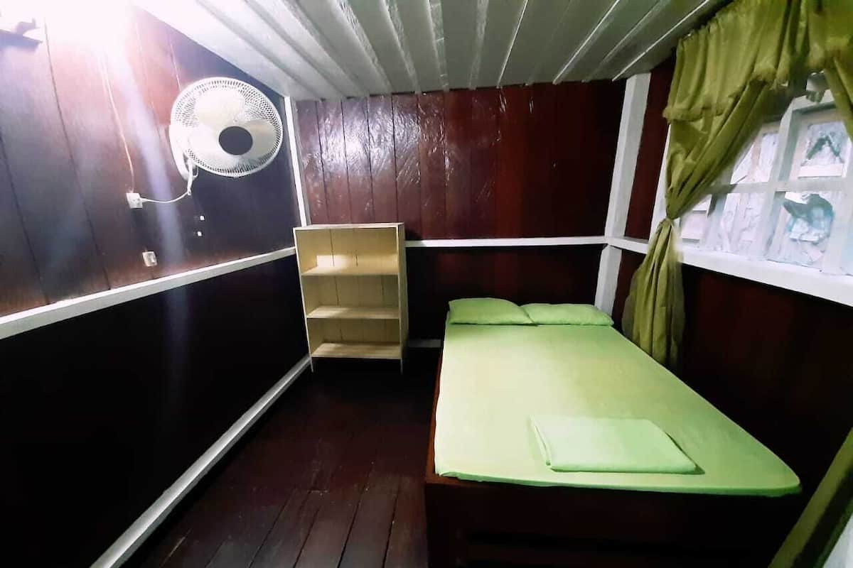 Une petite pièce, de taille modeste, comprenant un lit simple, une armoire en bois, un ventilateur fixé au mur et une fenêtre avec des rideaux.