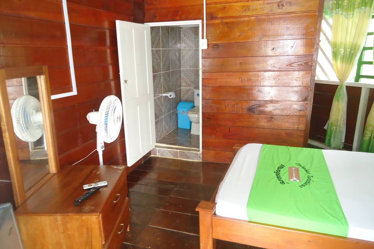 Une pièce en bois comprenant un lit, une commode, un miroir, un ventilateur et une salle de bain visible à travers une porte ouverte.