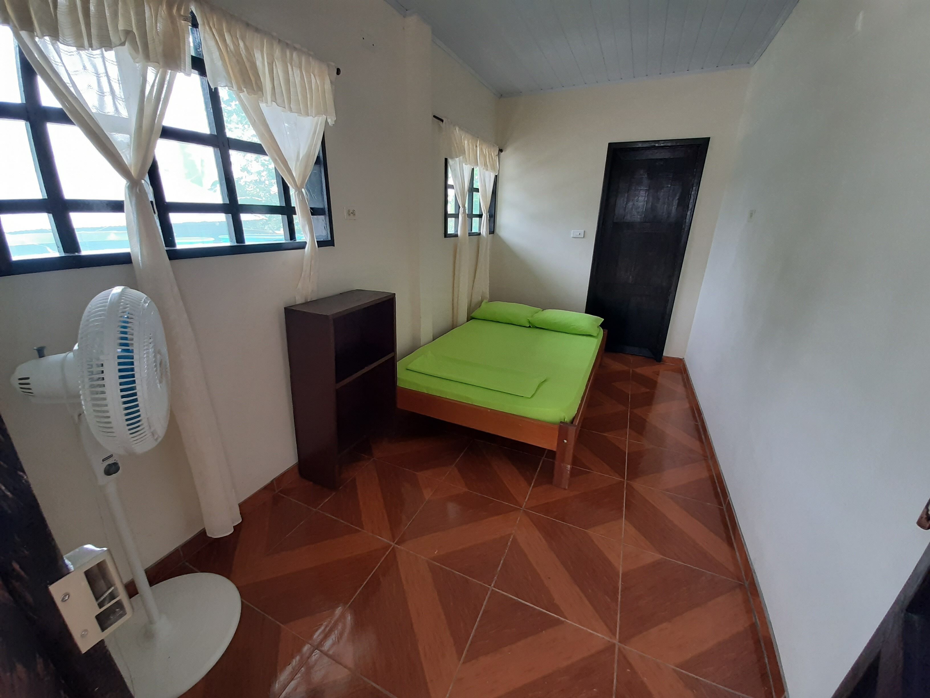 A room with a bed, a fan, a small cabinet, and a window with curtains.