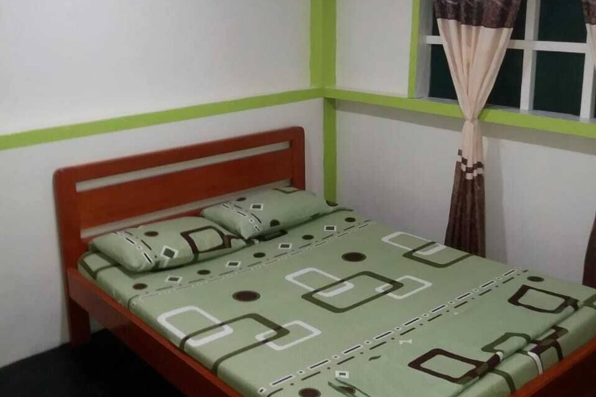 Un lit simple avec une couvre-lit à motifs verts et blancs, une tête de lit en bois et une fenêtre avec des rideaux.