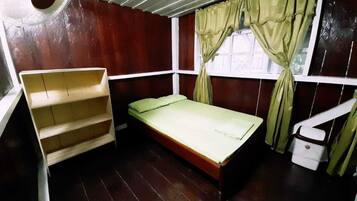 Una habitación pequeña y funcional con una cama individual, una estantería de madera y una ventana con cortinas.