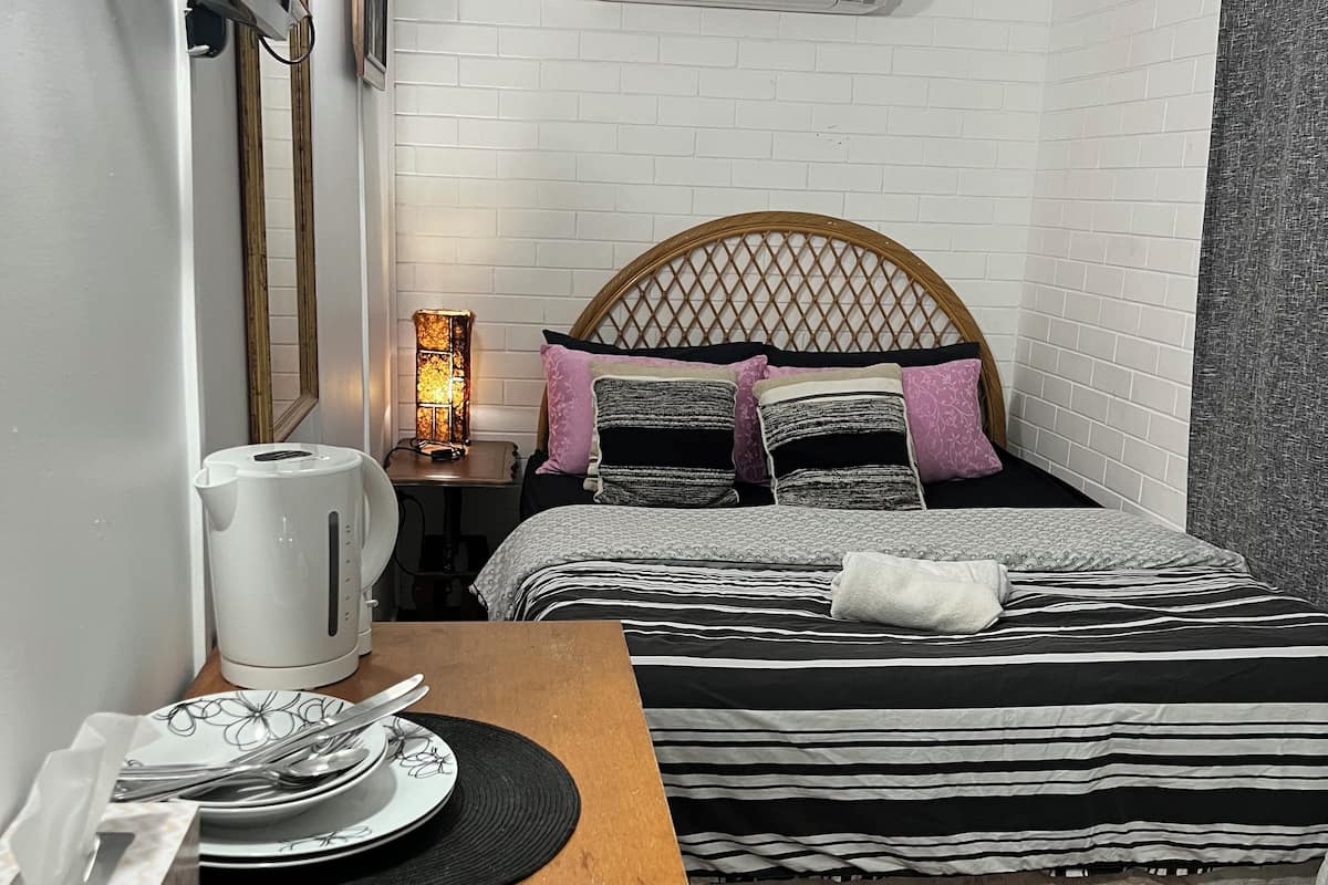 A hotel room with a bed, bedside table, lamp, and a small table with a kettle and plates.