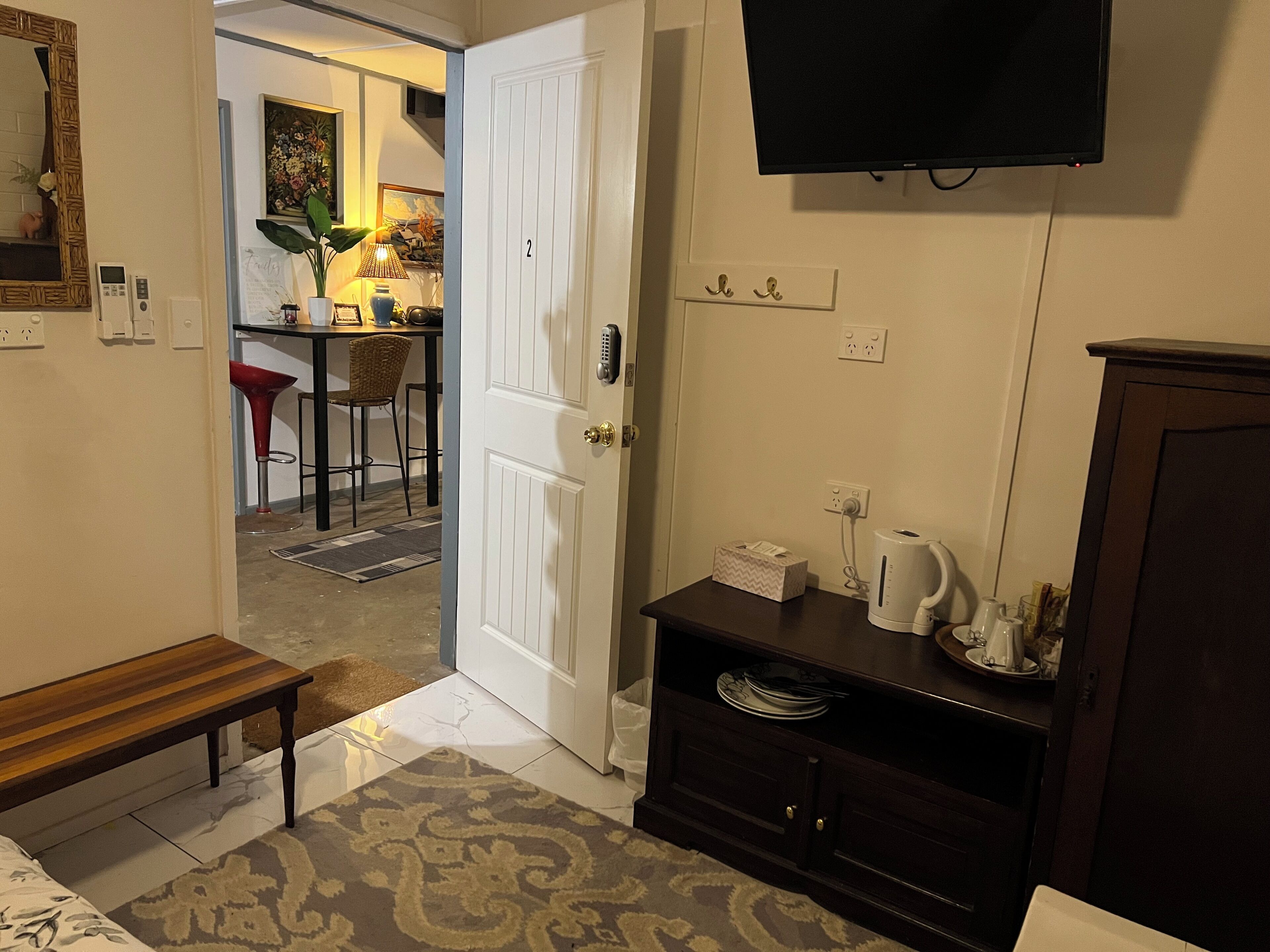 Une chambre d’hôtel avec un banc en bois, un tapis à motifs, une porte blanche, une petite table avec une bouilloire et un téléviseur fixé au mur.