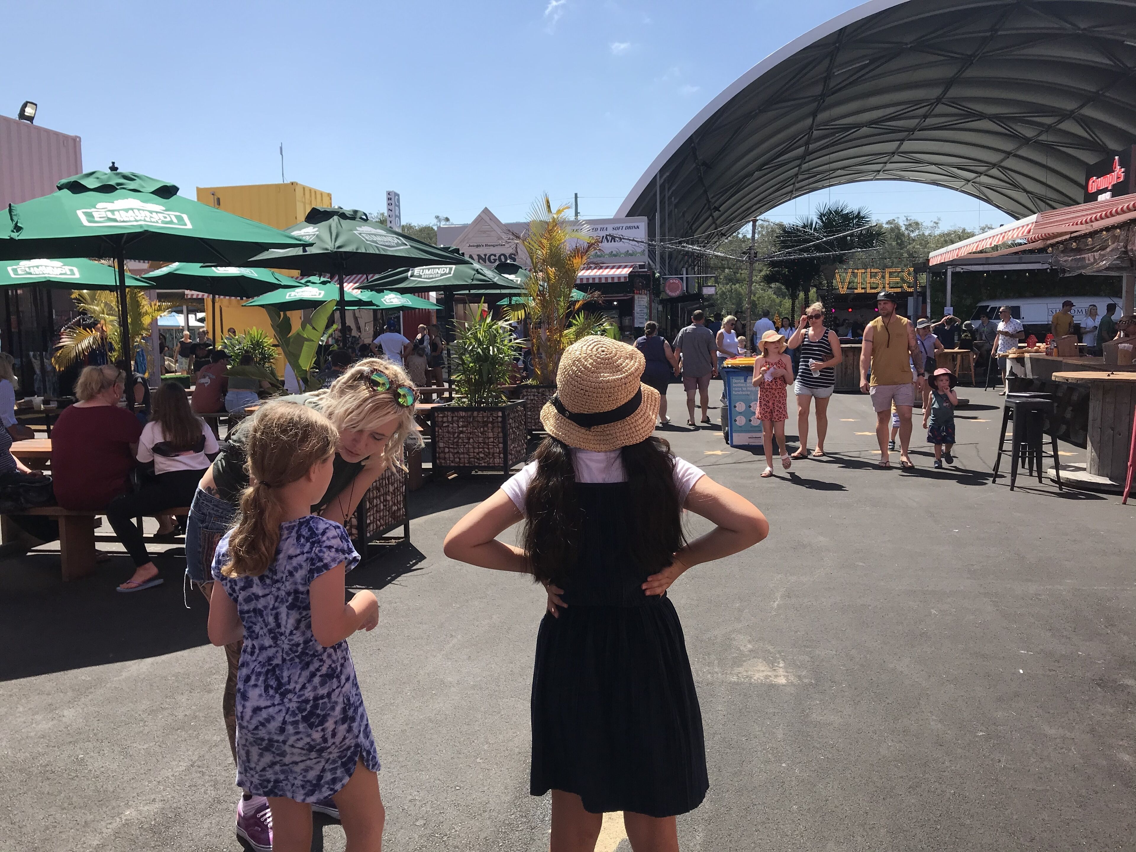 Un marché en plein air avec des parasols verts, des gens qui se promènent et des étals de nourriture.