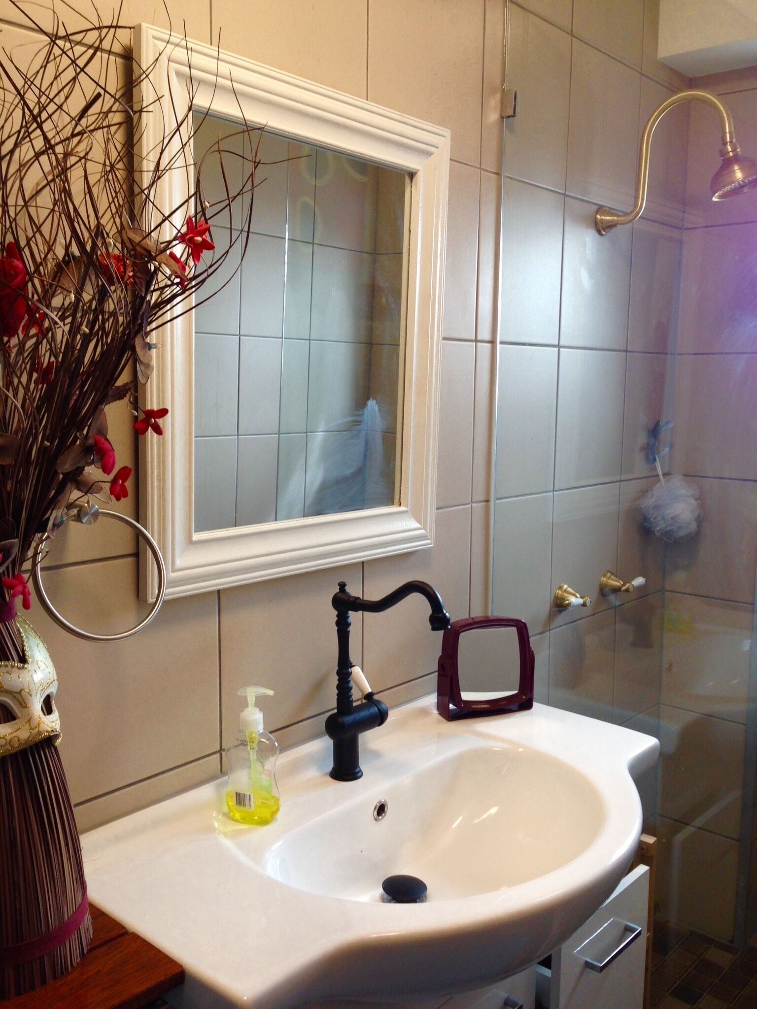 A bathroom with a white sink, a mirror, a shower, and a vase with dried branches and red flowers.