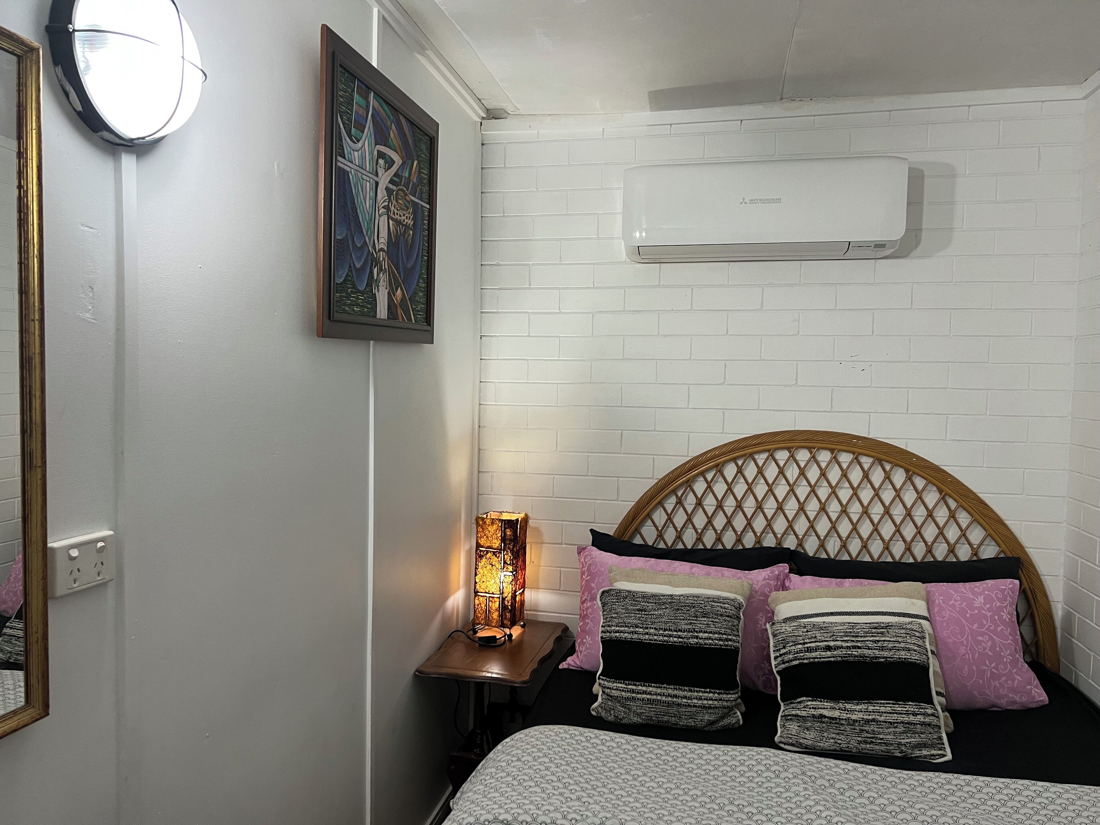 Un dormitorio con pared de ladrillo blanco, una cama con ropa de cama estampada, una mesita de noche con lámpara y un aire acondicionado de pared.
