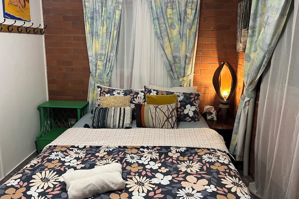 A bedroom with a floral bedspread, multiple pillows, a green nightstand, and a wall-mounted lamp.