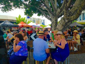 An outdoor dining area with tables and chairs under a large tree, surrounded by various food stalls.