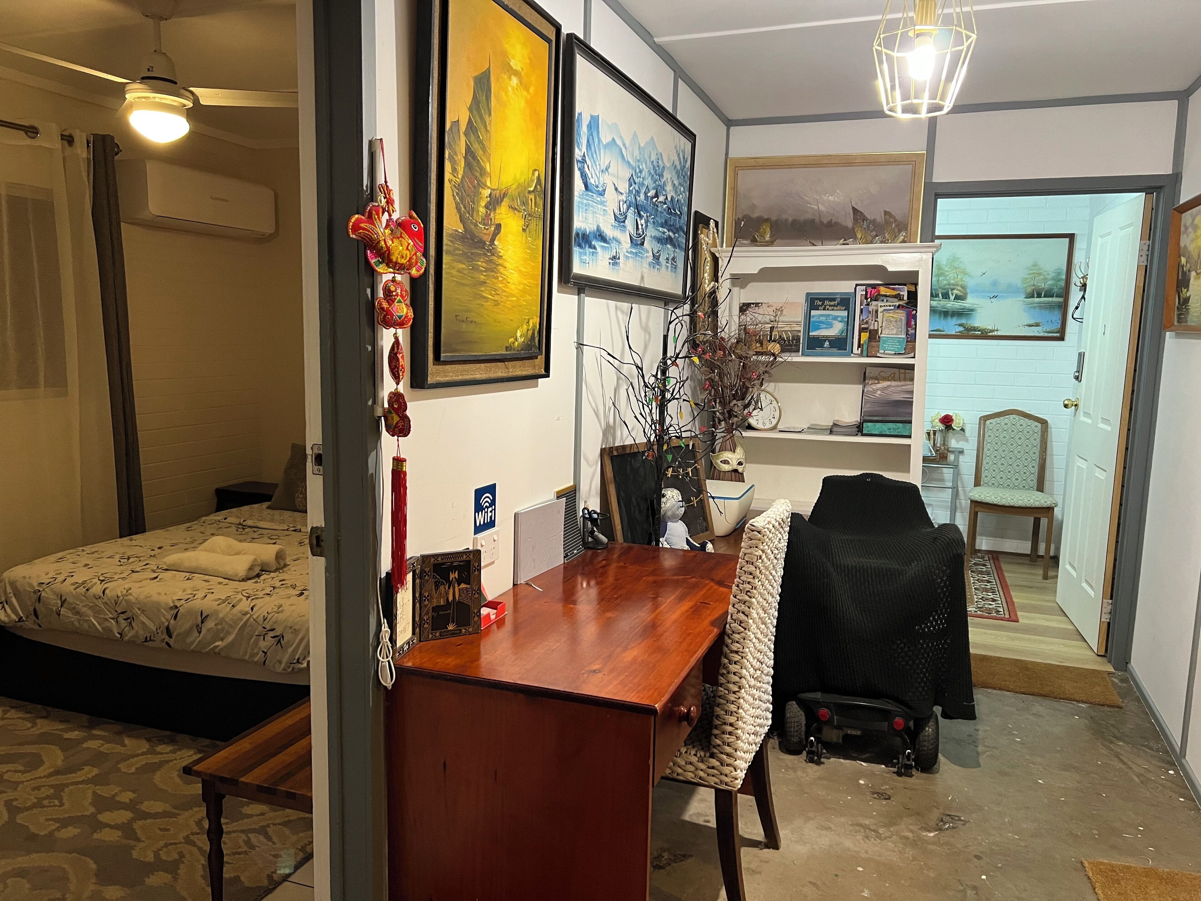 A room with a bed, a wooden desk, a chair, and a dining table. There are paintings on the walls and a ceiling fan.