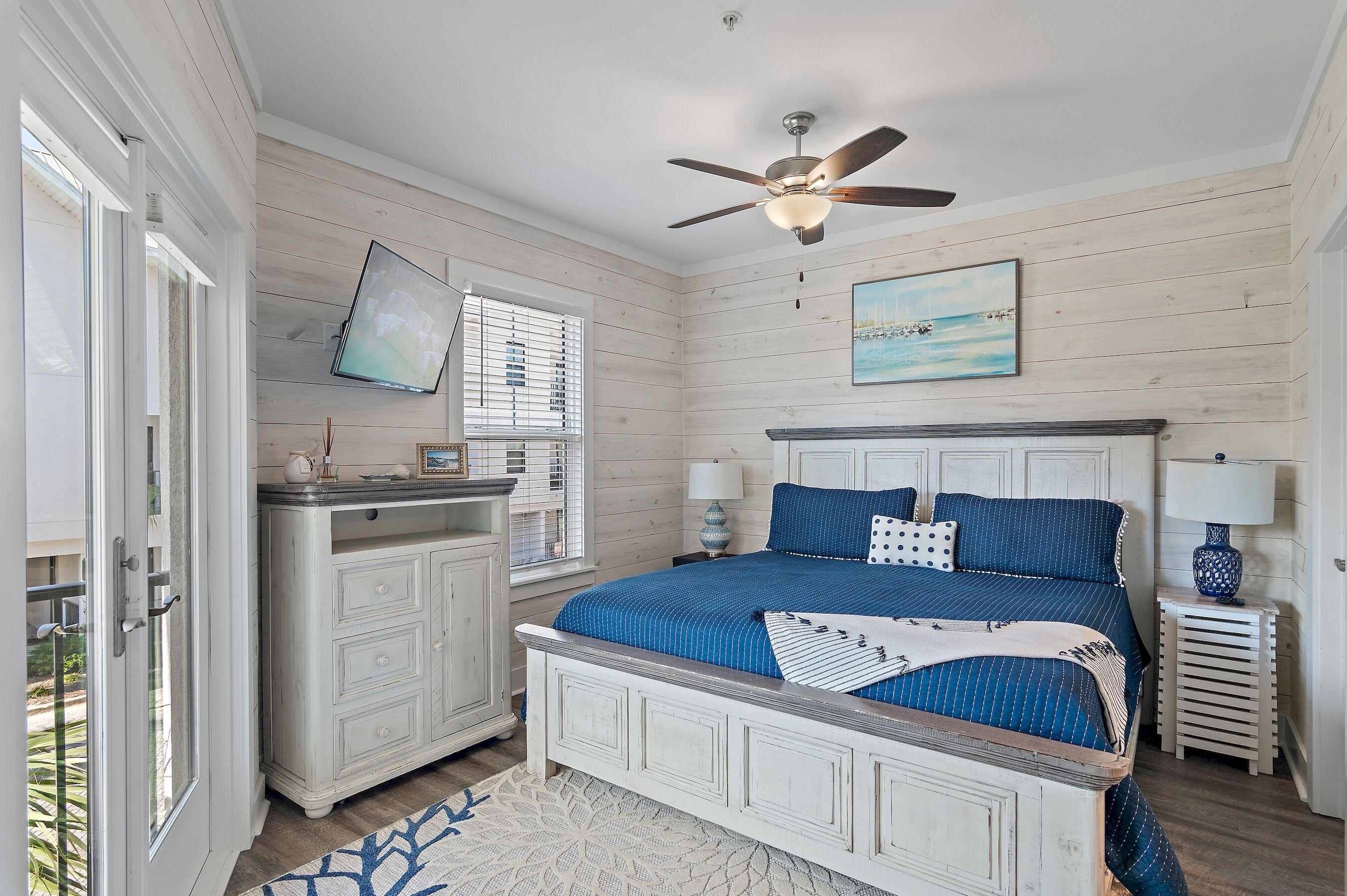 A bedroom with a bed, a ceiling fan, a TV, a desk, and a window with blinds.