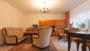 A living room with a sofa, armchairs, a coffee table, and a dining table with chairs. There is a TV, a small plant, and a window with curtains. - Pension Doemens (Aachen)