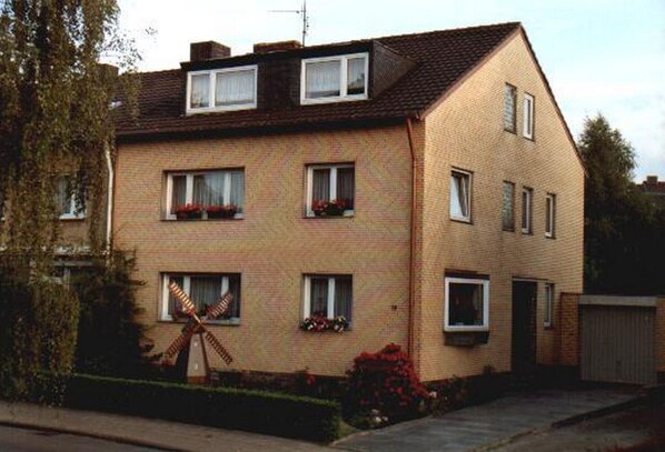 Un immeuble d’appartements en briques de deux étages, doté de nombreuses fenêtres, d’une antenne parabolique bien visible et d’un petit jardin agrémenté de fleurs.