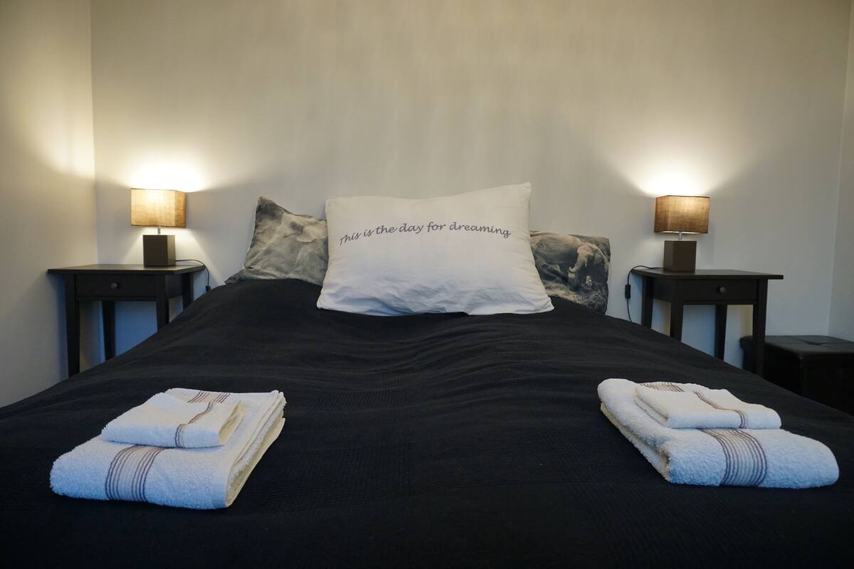 A neatly made bed with a pillow that reads "This is the day for dreaming," two bedside lamps, and folded towels on the bed.