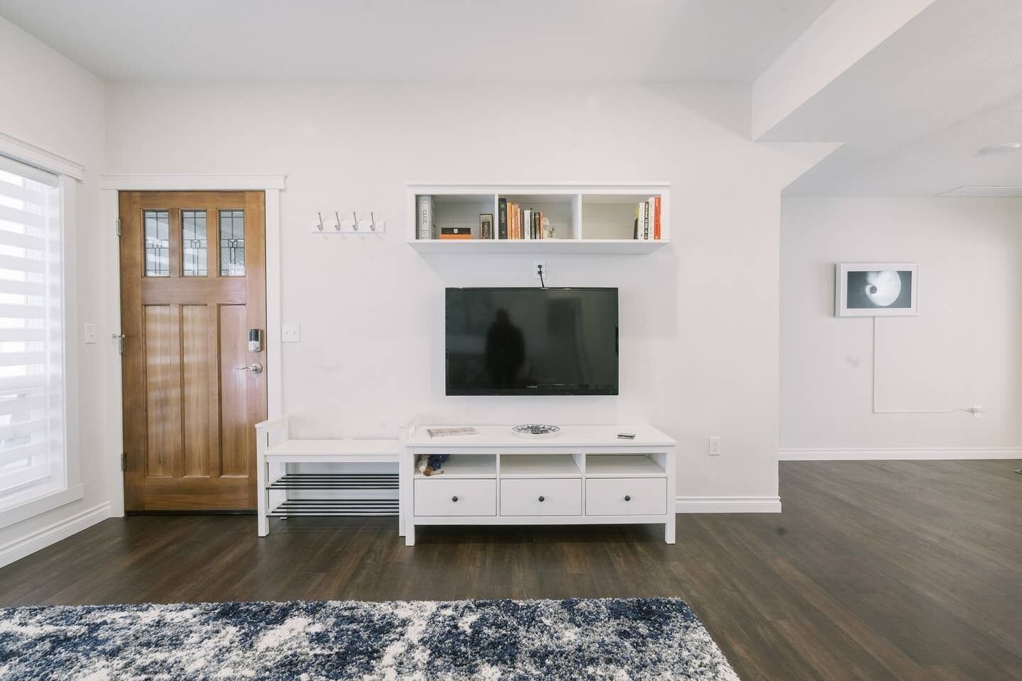 A modern living room with a wooden door, a white entertainment unit, a flat-screen TV, and a blue and white rug.