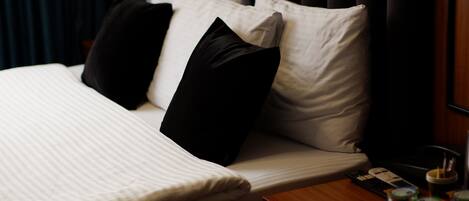 A neatly made bed with striped bedding, a wooden nightstand with a remote control, a tray with glasses, and a window with dark curtains.